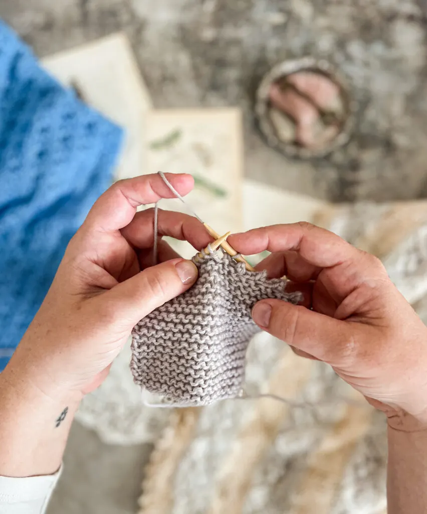 A white woman's hands (mine!) working the picot bind off on a gray garter-stitch square.