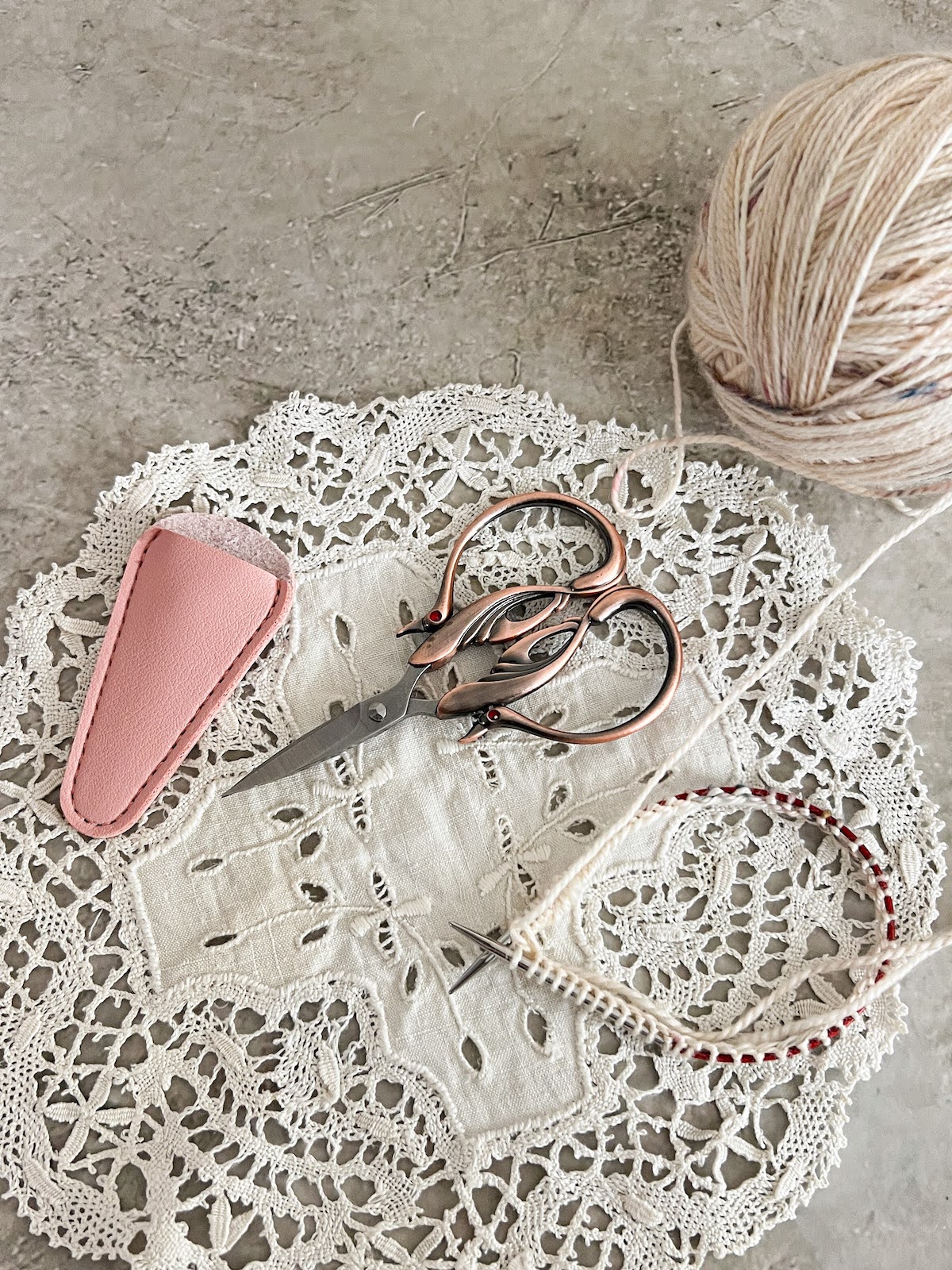 A pair of copper scissors with handles shaped like swans sits on a cream doily. On either side of the scissors are a pink sheath for the scissor blades and a tiny circular needle with the beginnings of a cream-colored sock on it.