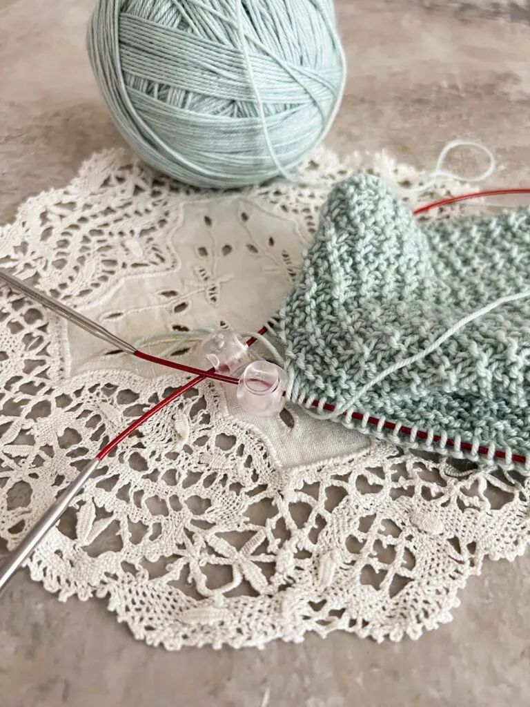 Two clear plastic cable locks are slid onto the red cable between two circular needles, one at each end. They are holding a seafoam green shawl in progress on the needles and preventing the needles from slipping out.