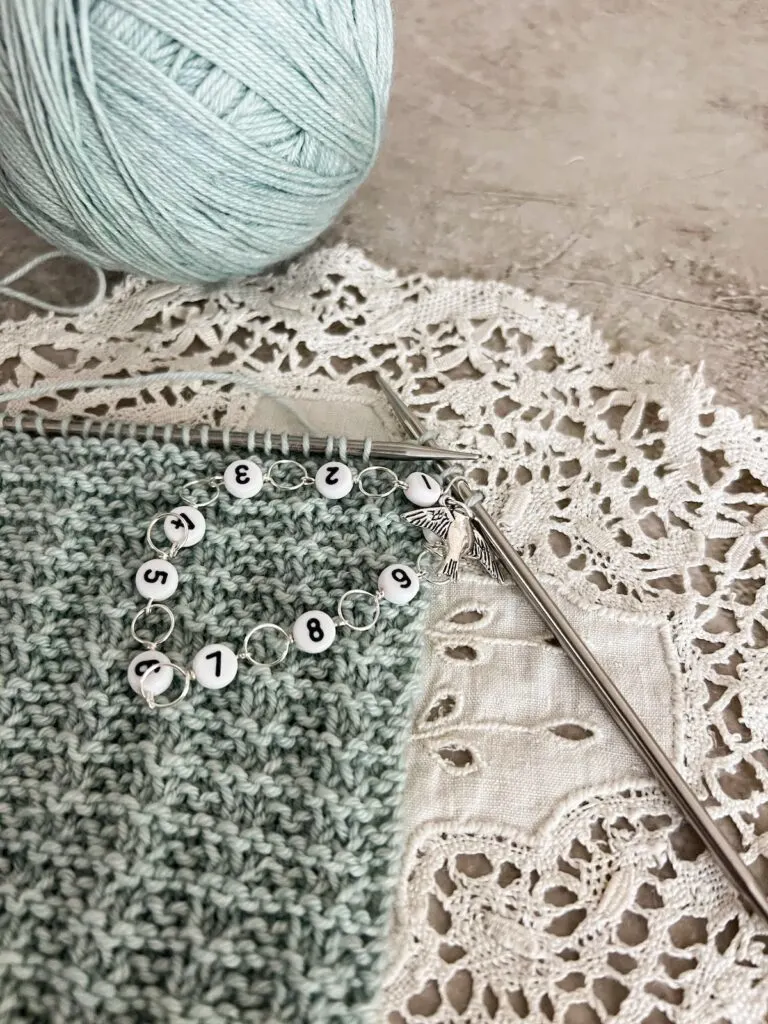 A row counter made of loops and numbered beads dangles from the needles of a seafoam green shawl in progress. The row counter also has a silver bird charm.