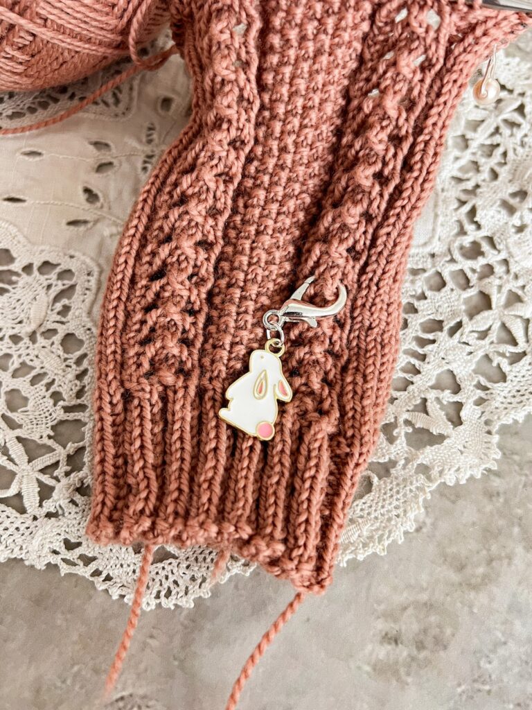 A pink and white enamel bunny progress keeper is clipped onto a pink sock in progress to track how many repeats of the stitch pattern have been completed.