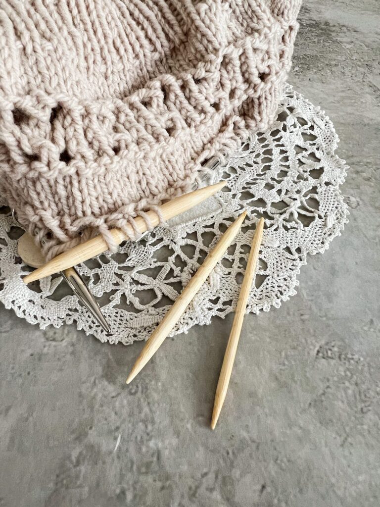 Three wooden cable needles in three different sizes. The needles are straight with points at each end and have grooves in the middle to hold stitches in place. The largest needle is shown in use holding some stitches on a tan hat in progress.