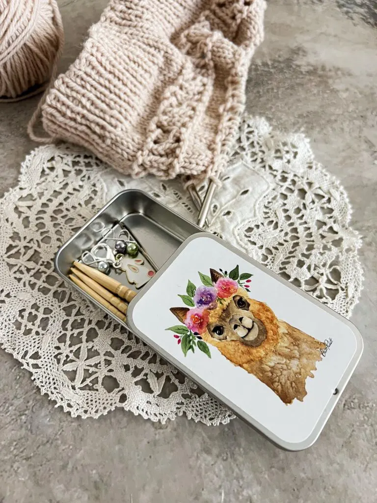 A metal notions tin with a drawing of an alpaca on the front is half-opened on a tabletop. Inside are stitch markers, darning needles, and cable needles. The alpaca on the tin is wearing a flower crown.