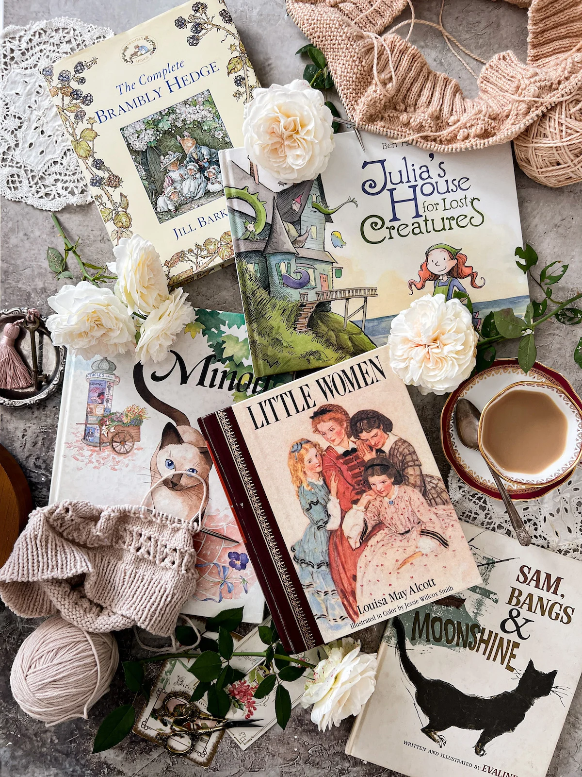 Five children's books are laid out flat on a gray surface, surrounded by several white roses, some knitting projects, a teacup and saucer, and a couple antique doilies. The books are The Complete Brambly Hedge, Julia's House for Lost Creatures, Minou, Little Women, and Sam, Bangs, and Moonshine.