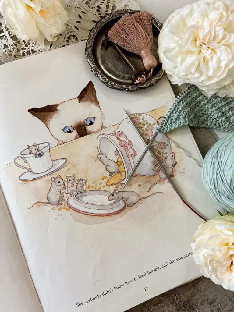 A flatlay showing a page from the inside of Minou. It shows a little Siamese cat with bright blue eyes peering over the edge of a countertop to watch some mice stealing nibbles of biscuits and tea.
