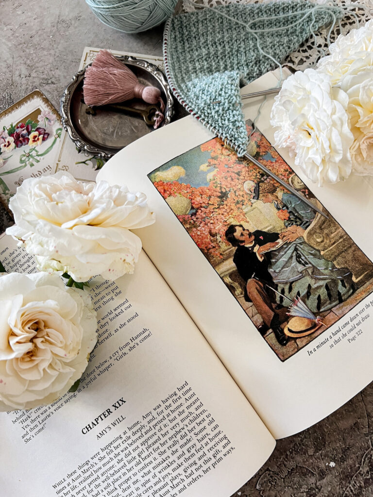 A flat-lay showing a page from inside Little Women, where a dashing young man with dark hair and a moustache kneels at the feet of a young blond woman in a light blue dress. Amy is trying to draw, but Laurie has placed his hand over the page to get her attention.