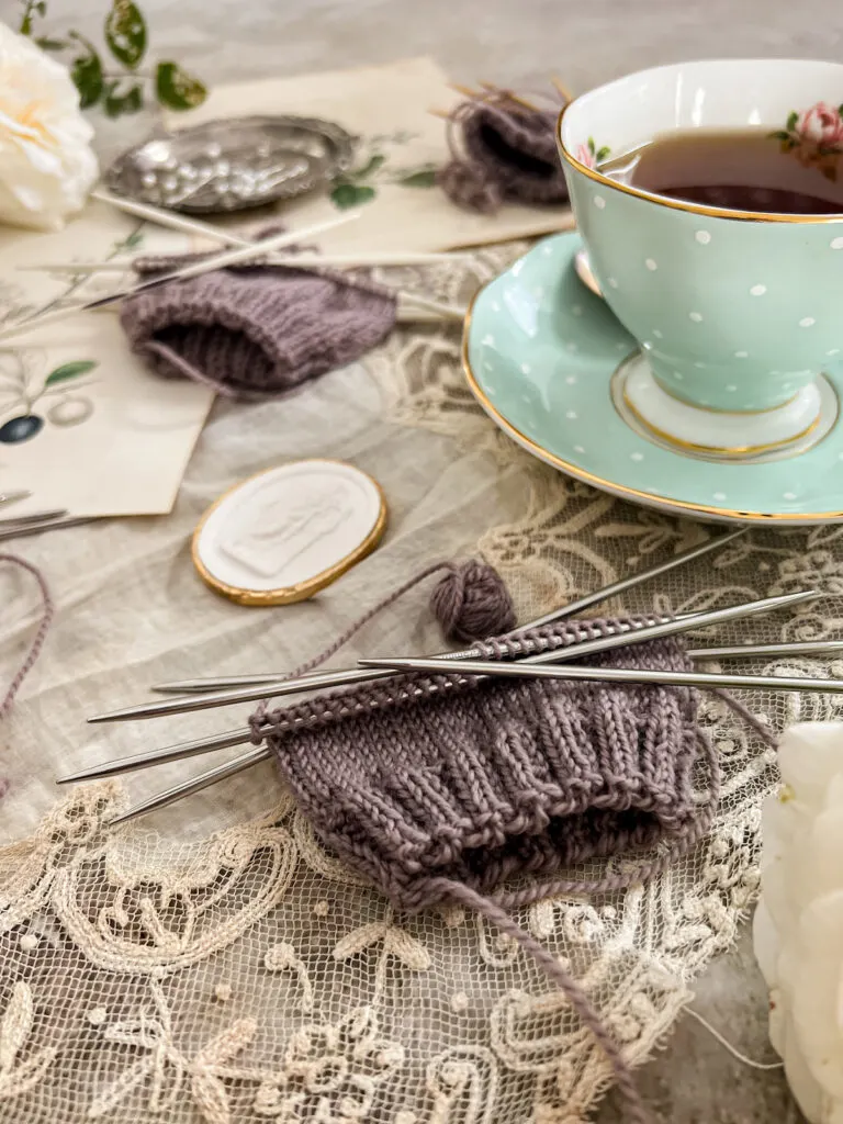 A close-up on a purple swatch knit using ChiaoGoo steel double-pointed needles. Slightly blurred in the background are a small plaster intaglio, other purple swatches on other needles, and a mint-colored teacup filled with tea.