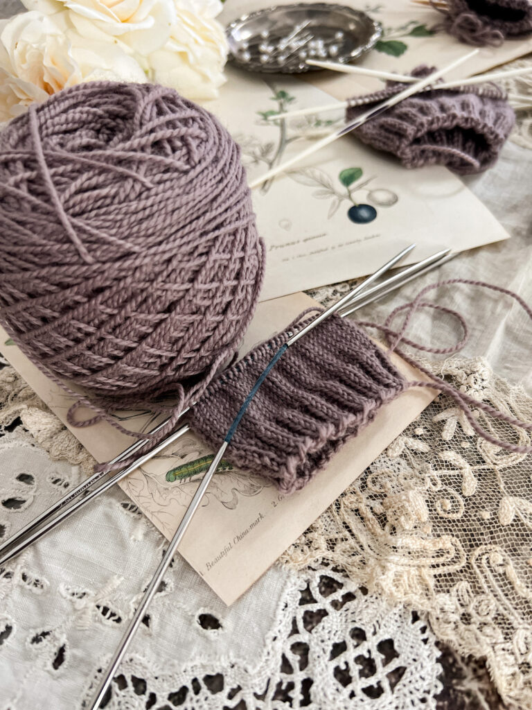 A close-up image showing a purple swatch knit on Addi FlexiFlips, a type of double-pointed needle with a bendable center so you can work with fewer needles than when you use traditional DPNs. The swatch is centered in the middle of the photo. In the backaground, blurred, are another purple swatch and some white roses.