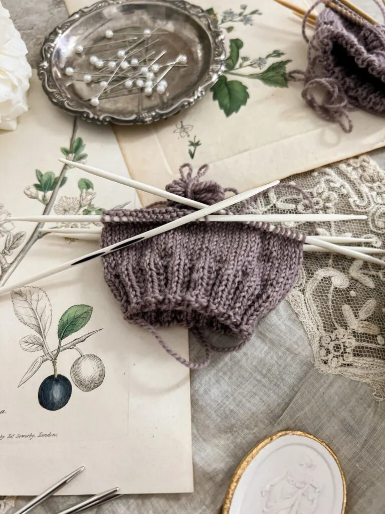 A close-up image showing a purple swatch knit on a set of Prym Ergonomics, a type of plastic double-pointed needles designed to help knitters who experience hand pain while working. One of the needles is laid across the top of the swatch so you can see its shape and markings. The swatch is on top of antique paper ephemera and textiles. There is a small plaster intaglio in the foreground and a tiny silver tray full of sewing pins in the background.