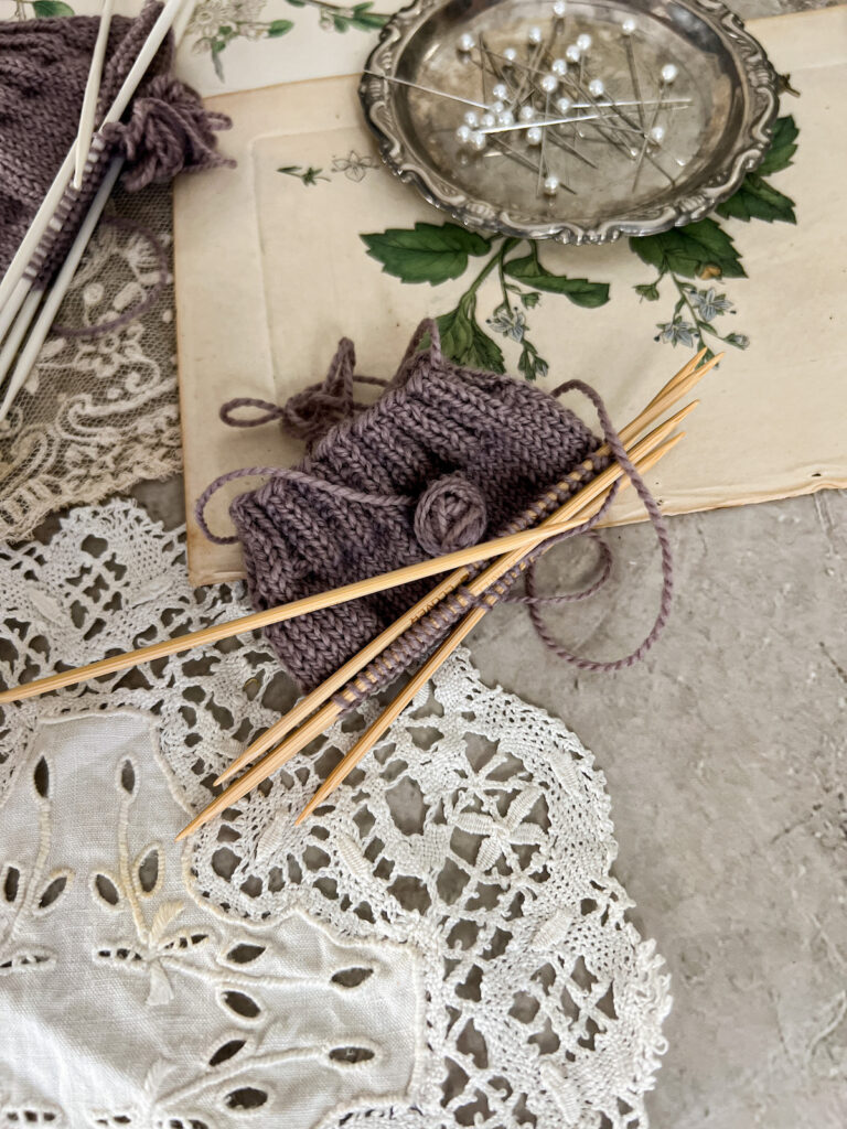 A close-up on a swatch knit with bamboo double-pointed needles. This purple swatch is surrounded by antique paper ephemera and textiles, along with a tiny silver tray filled with sewing pins (blurred in the background).