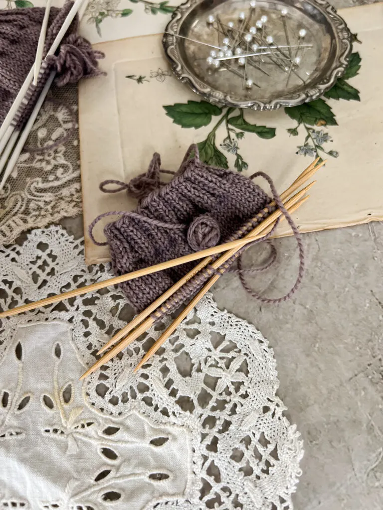 A close-up on a swatch knit with bamboo double-pointed needles. This purple swatch is surrounded by antique paper ephemera and textiles, along with a tiny silver tray filled with sewing pins (blurred in the background).