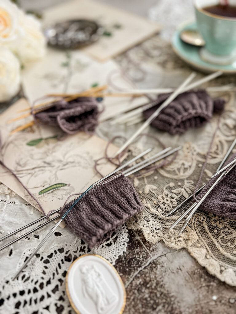 A photo of four swatches knit in different kinds of double pointed needles. The photo is taken from a low angle, and only the lower left quadrant is in focus. It shows a purple swatch knit on Addi FlexiFlips.