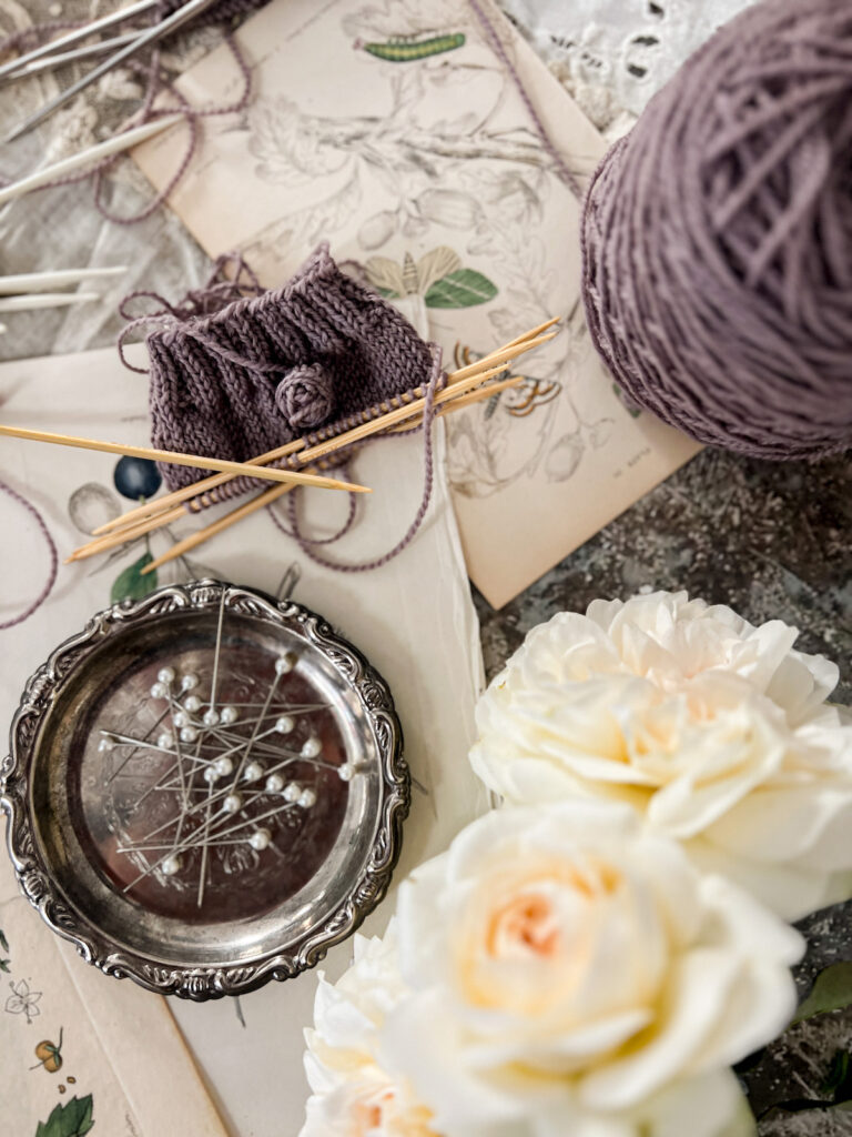 A top-down photo showing a purple swatch knit on a set of Takumi Clover bamboo double-pointed needles. The swatch is in the top left corner of the image, and it sits on top of antique botanical illustrations. It's surrounded by a tiny silver tray filled with sewing pins, a purple cake of yarn, and a blurry bunch of white roses.