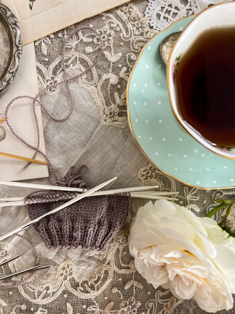 A top-down image of a purple swatch knit using Prym Ergonomics, a type of triangular-shaped double-pointed needle. The swatch is in the bottom left of the photo. A mint-colored cup of tea fills the top right.