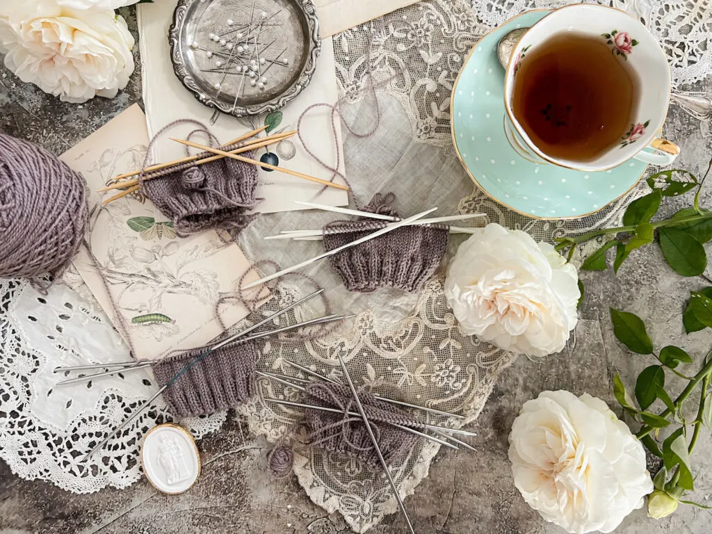 A flatlay showing four purple swatches on four different kinds of double-pointed needles. They're surrounded by antique textiles and paper ephemera, white roses, and a mint-colored teacup full of tea.