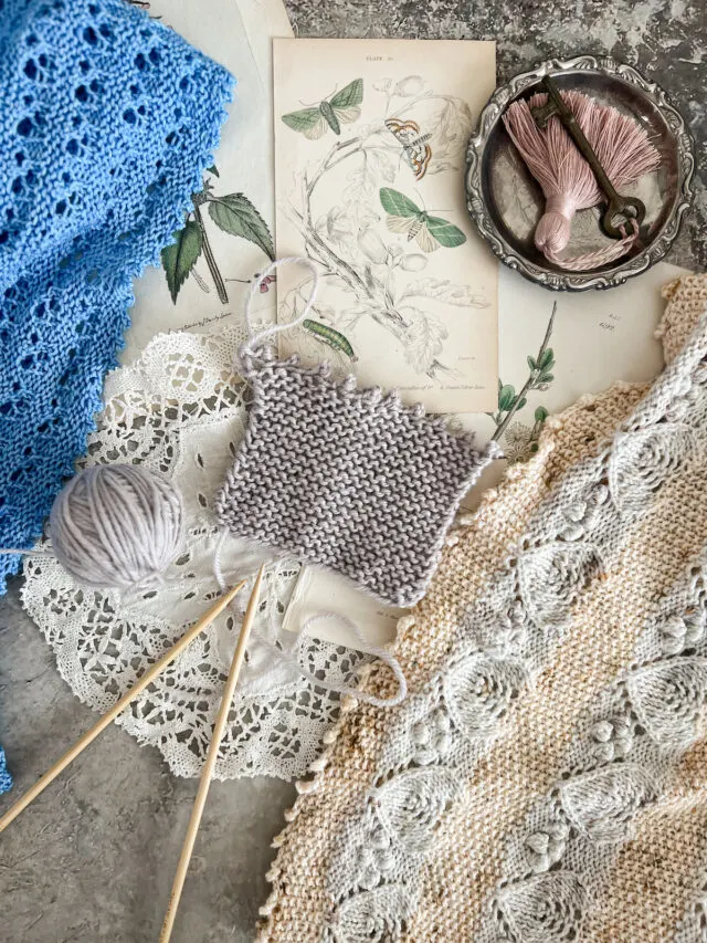 A top-down photo showing a gray garter-stitch square with a picot bind off. To the left is a blue shawl with a picot bind off. To the right is a yellow and blue shawl with a picot bind off. There are also antique botanical prints, wooden knitting needles, a doily, and a small silver tray with a brass key that has a tassel tied to it.