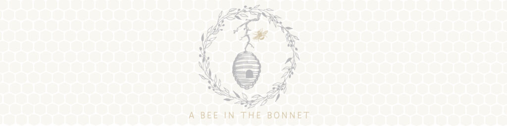 A banner image with a honeycomb background in a golden color and a gray beehive surrounded by a wreath. A single golden bee flies toward the opening of the hive. Underneath the hive are the words A Bee In The Bonnet.