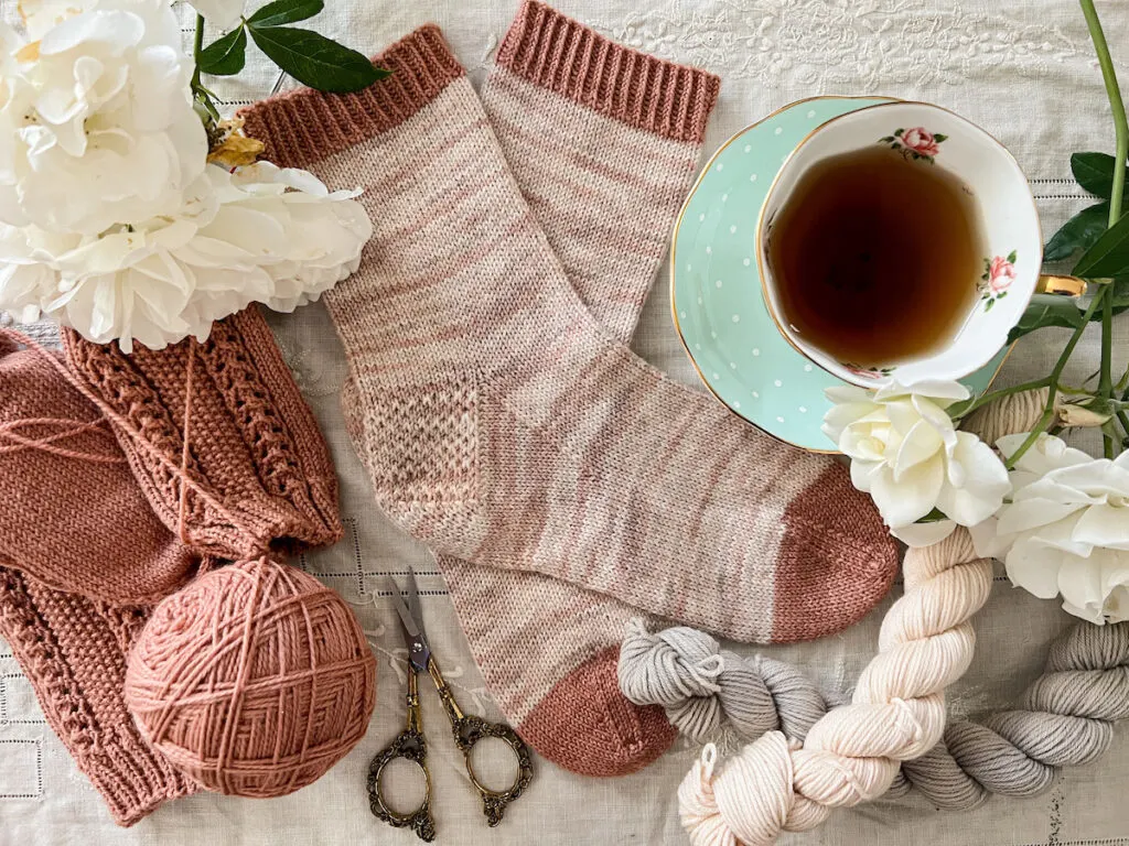 A top-down photo of a pair of light pink vanilla socks laid flat on a table with the toes pointing to the right. One sock is crossed over the other. They're surrounded by white roses, a few mini skeins of neutral yarn, a pair of dark pink socks in progress, and a mint-colored teacup full of tea.