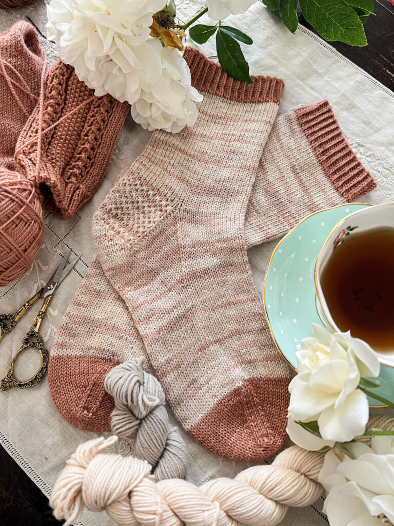 A side-angle photo of a pair of light pink vanilla socks laid flat on a table top. They're surrounded by white roses, a few mini skeins of neutral yarn, a pair of dark pink socks in progress, and a mint-colored teacup full of tea.