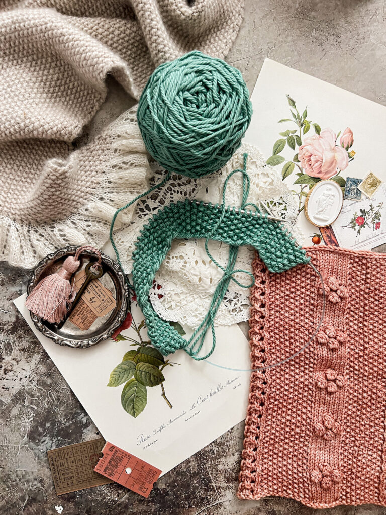 A vertical flatlay photo showing a gray seed stitch shawl (top left), a turquoise seed stitch dishcloth in progress (center), and a pink cowl with seed stitch panels (bottom right). They're surrounded by antique paper ephemera and trinkets.