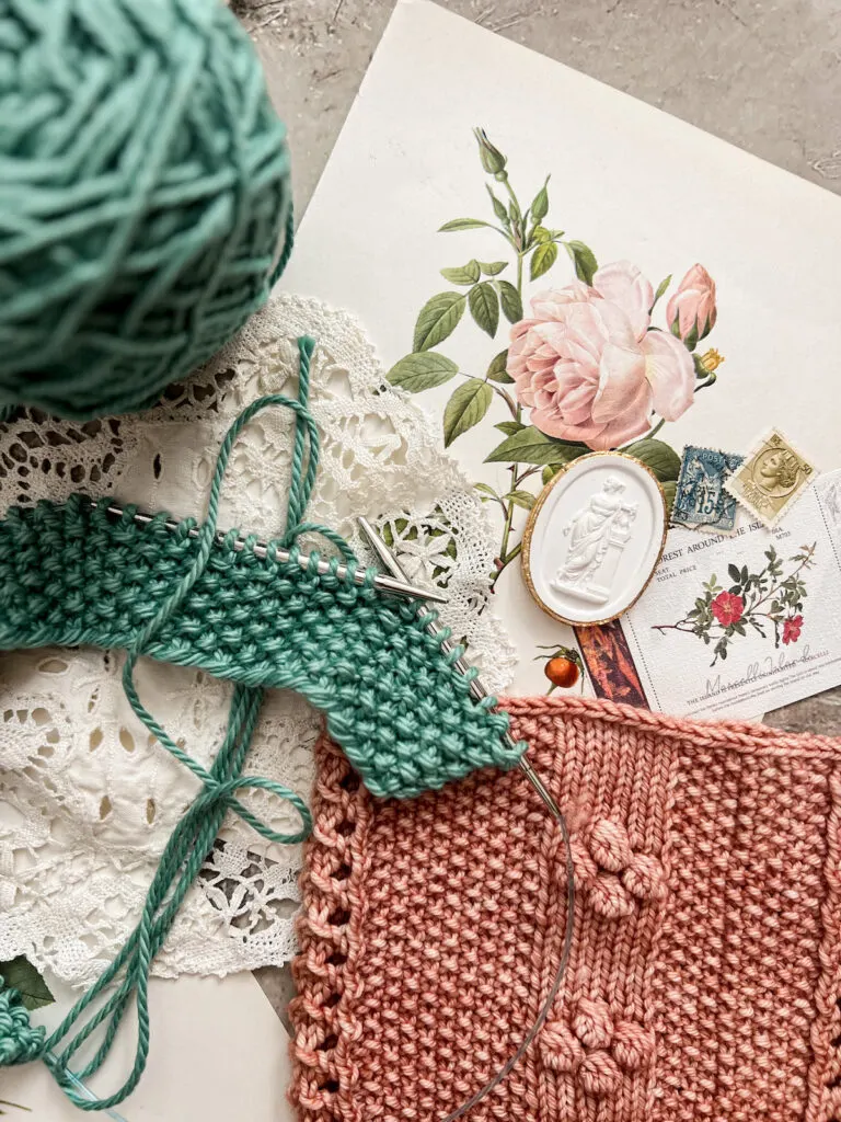 A top-down image showing a tuquoise dishcloth in progress laid on top of a pink cowl with seed stitch panels. They're surrounded by antique paper ephemera and a white intaglio.