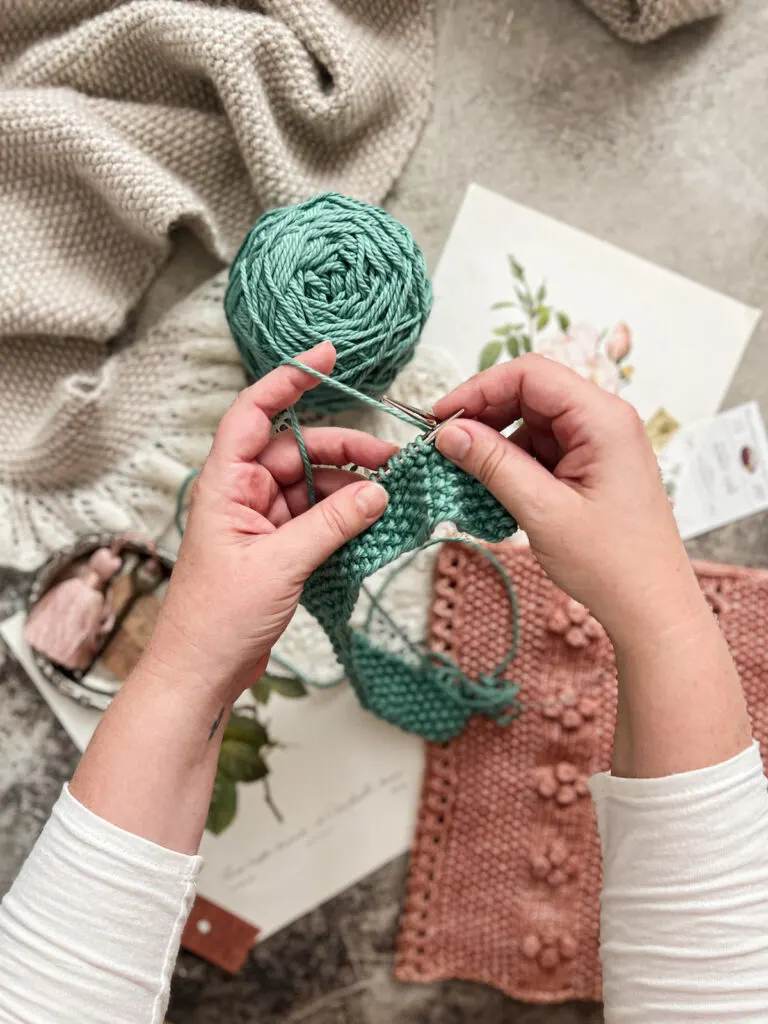 A white woman's hands (mine!) work on a turquoise dishcloth. The dishcloth is knit in seed stitch.