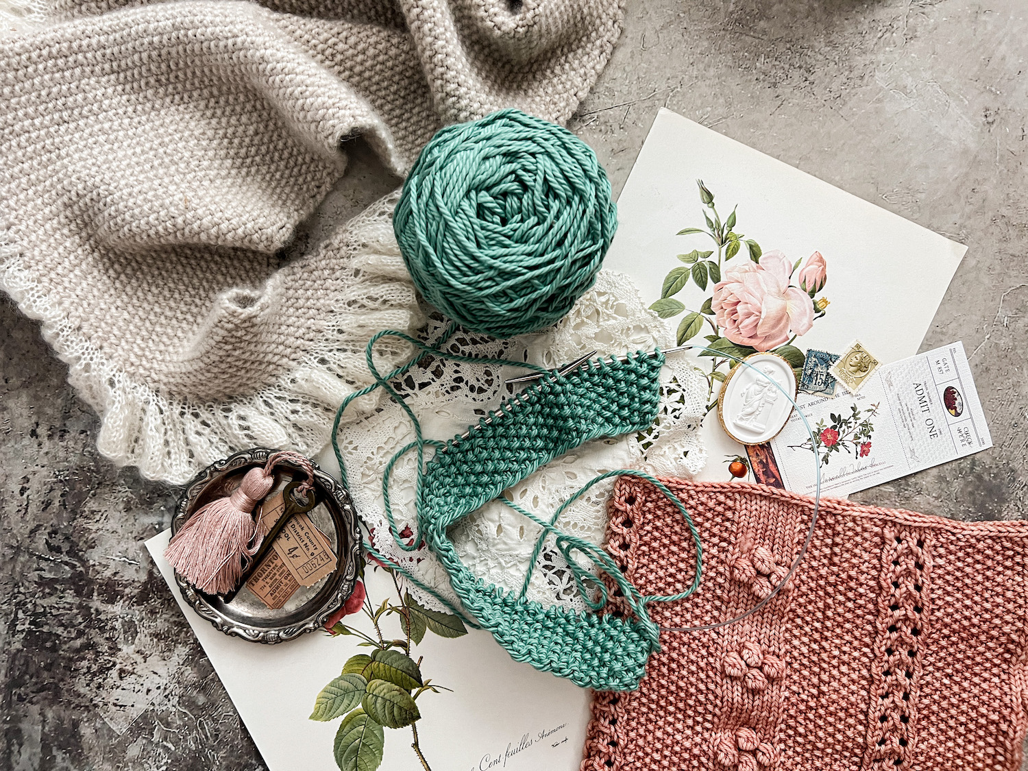 A horizontal photo showing three projects made with seed stitch: a gray shawl at top left, a pink cowl at bottom right, and a turquoise dishcloth in progress in the middle. they're surrounded by antique paper ephemera and trinkets.