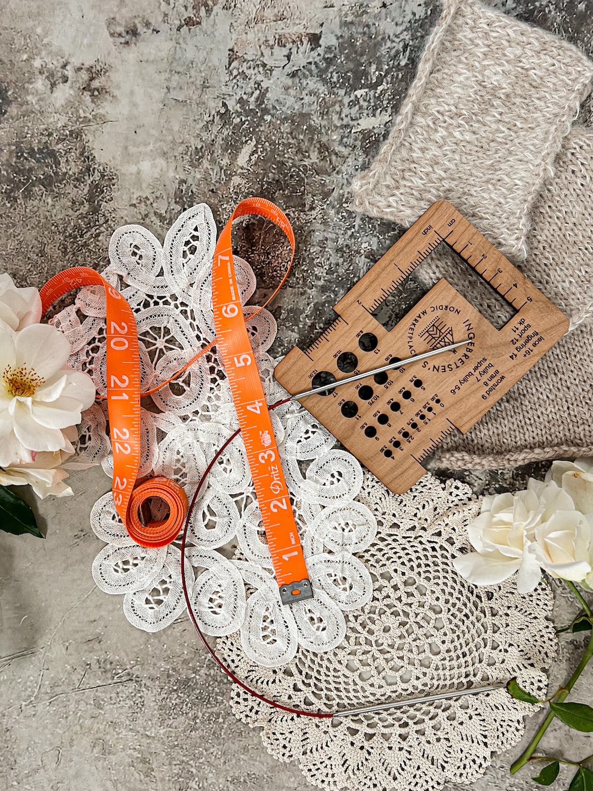 A top-down photograph showing an orange tape measure, a circular knitting needle, and a wooden needle gauge finder. They are surrounded by white roses, a few gauge swatches, and some antique doilies.
