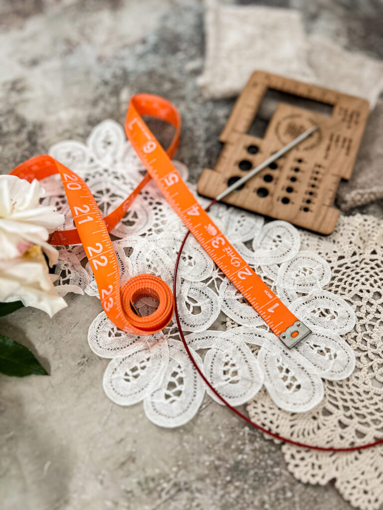 A side-angle photograph showing an orange tape measure and part of a circular knitting needle. Blurred in the background is a wooden needle gauge finder.