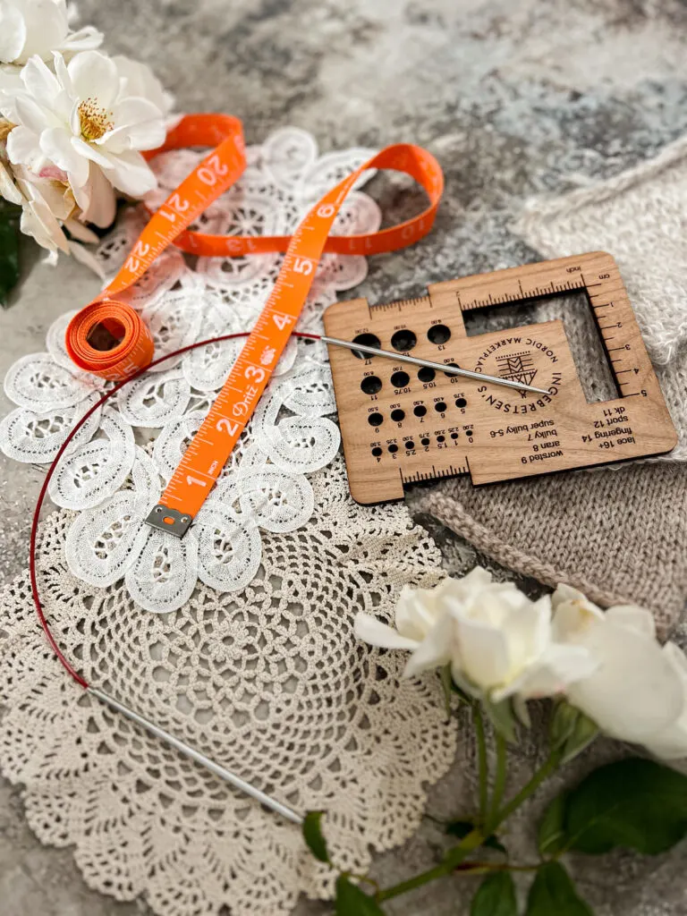 A side-angle photo of a circular knitting needle on a tan antique doily. Slightly blurred in the background are a wooden needle gauge finder and an orange tape measure.