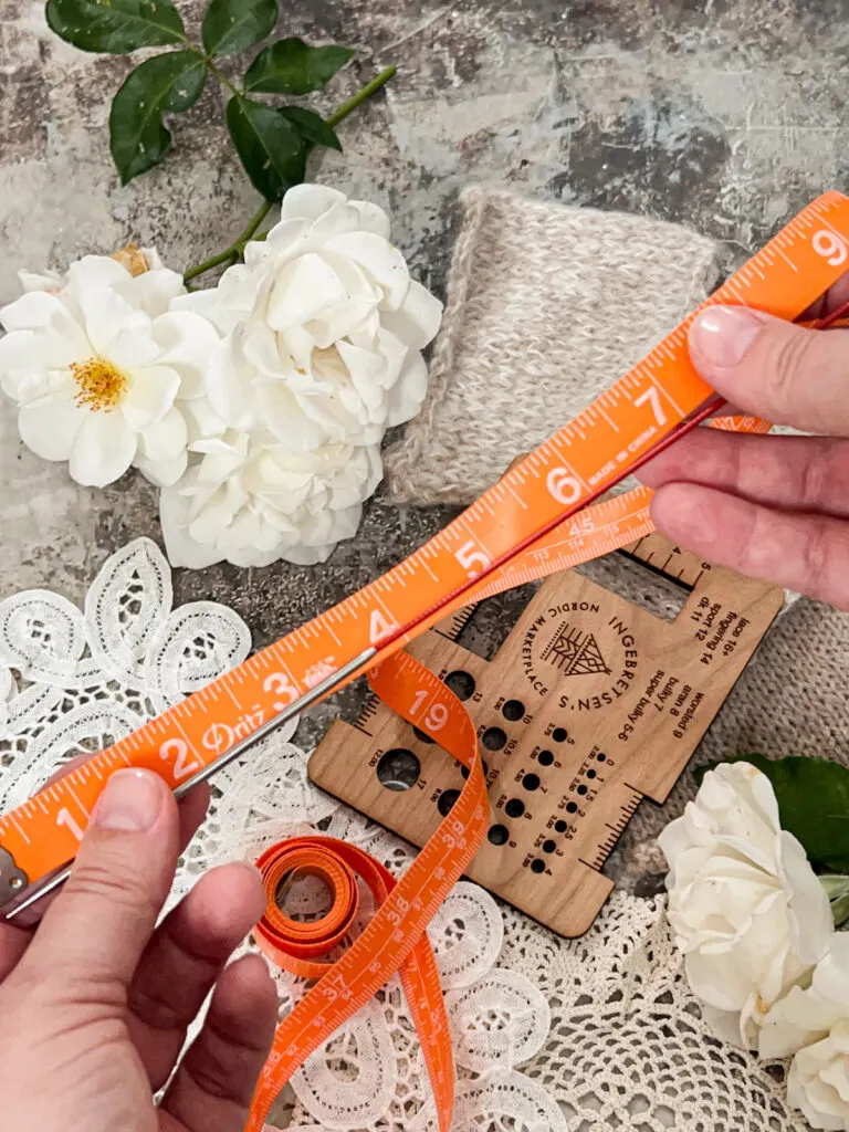 A white woman holds an orange measuring tape and a circular knitting needle to see how long the needle is.