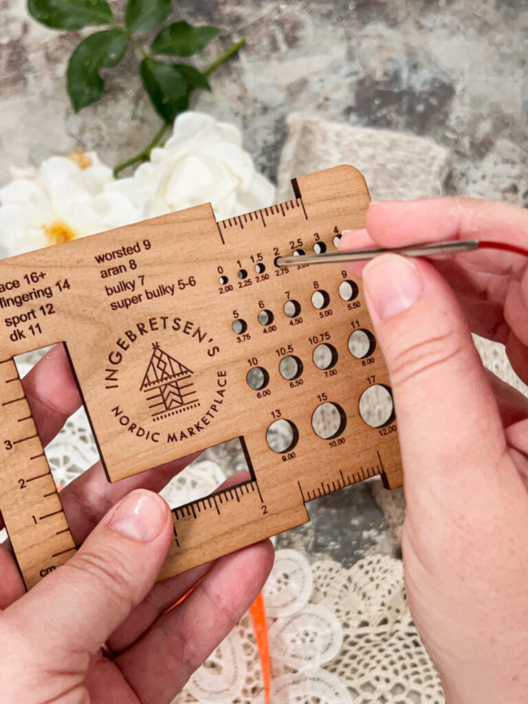 A white woman's hands hold a wooden needle gauge finder while inserting a knitting needle into one of the measuring holes.