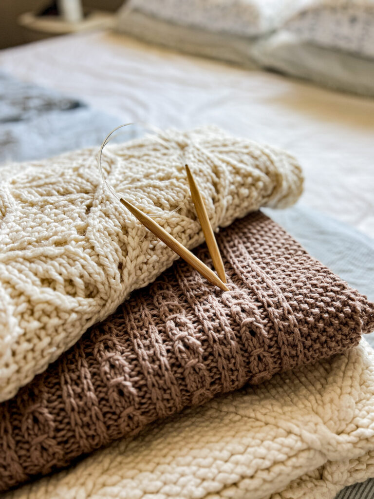 A side-angle photo of the three stacked knit blankets and the pair of knitting needles. Blurred in the background is the rest of the neatly made blue-and-white bed they're resting on.