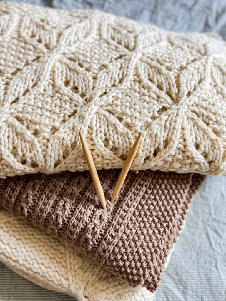 Another angle of the pile of three knit blankets and wooden circular needles. The circular needles are in the center of the photo. The blankets are cream (top), brown (middle), and cream (bottom).
