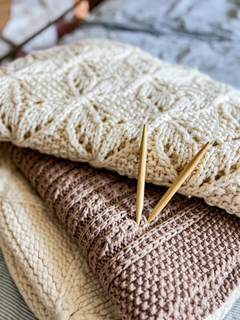 A pile of three knit blankets is stacked crookedly so all three blankets are visible. A pair of wooden circular needles rests on top of the stack.