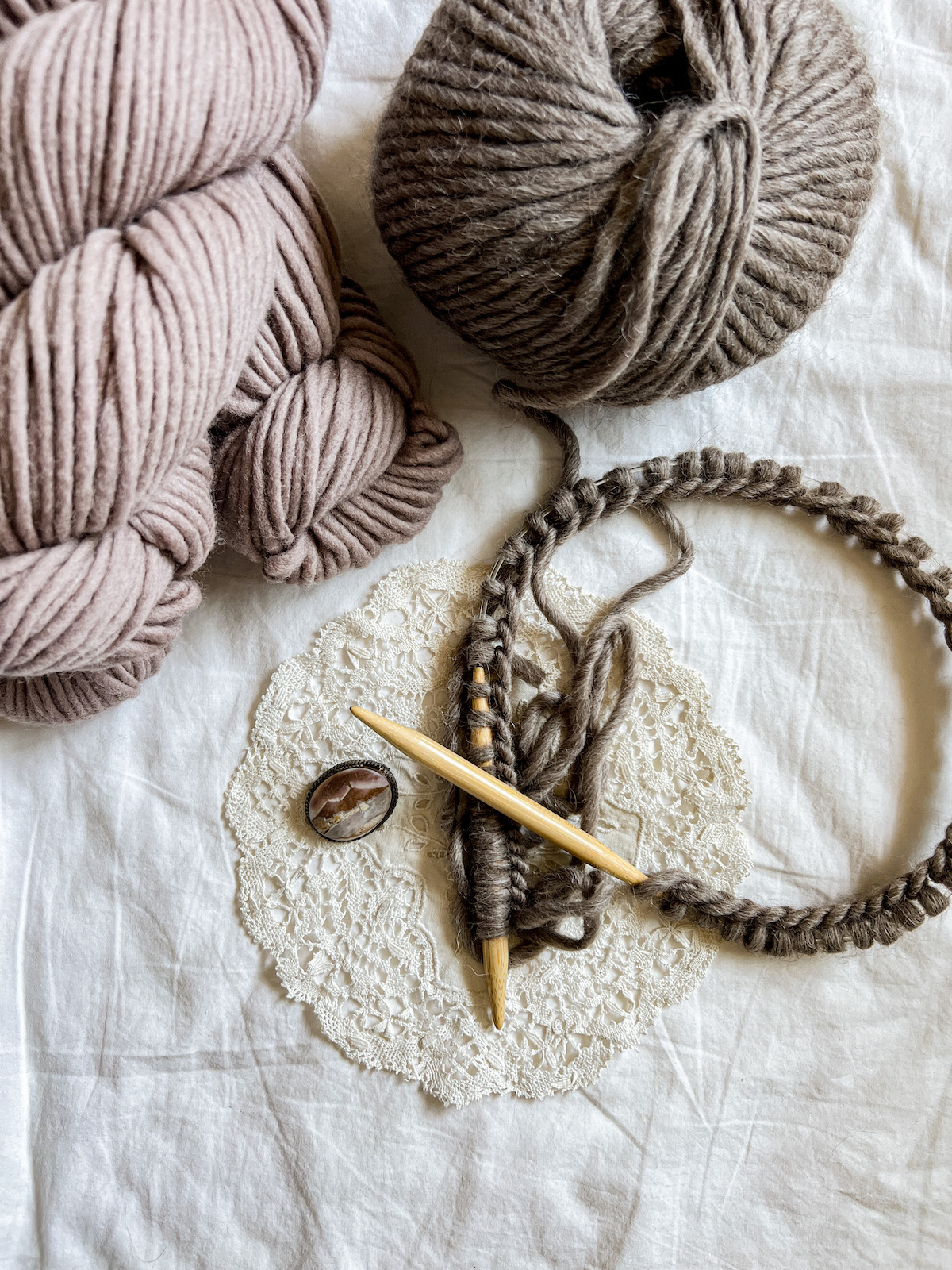 A flatlay photo of a few skeins of medium-brown yarn, a ball of dark brown yarn, and some wooden circular needles with lots of dark brown stitches cast on to them. The needles are resting on a white lace antique doily with a carved shell brooch next to them.