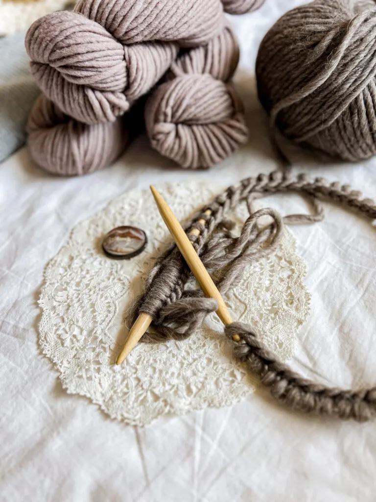 In the foreground is a pair of wooden needles with lots of dark brown stitches cast on. They rest on a white antique doily. In the background are several skeins and a ball of brown yarn.