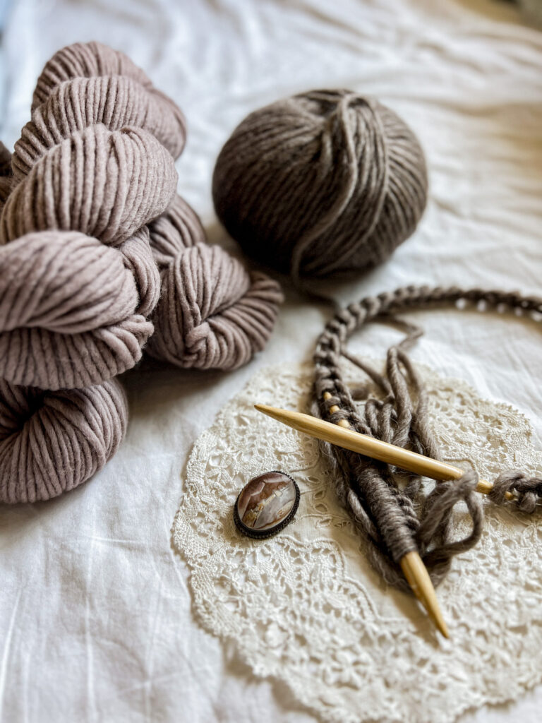 A side-angle photo of a few skeins of brown yarn, a ball of darker brown yarn, wooden circular needles with lots of dark brown stitches cast on, and a small carved shell brooch.
