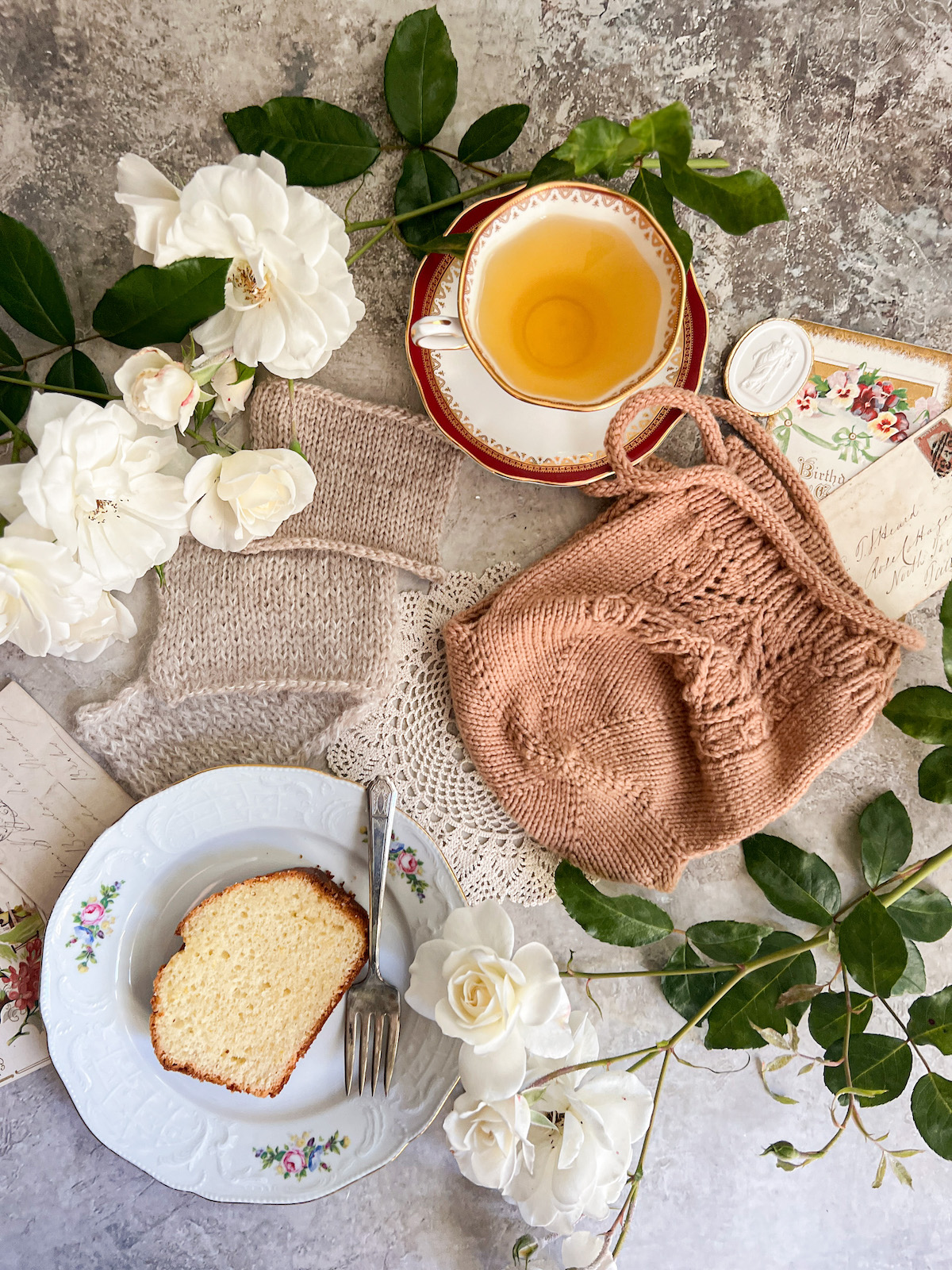 A flatlay photo showing a tea cozy with lots of KFB increases on its bottom, some antique paper ephemera, white roses, a vintage doily, a plate with a slice of cake, three knitting swatches, and a teacup full of chamomile.