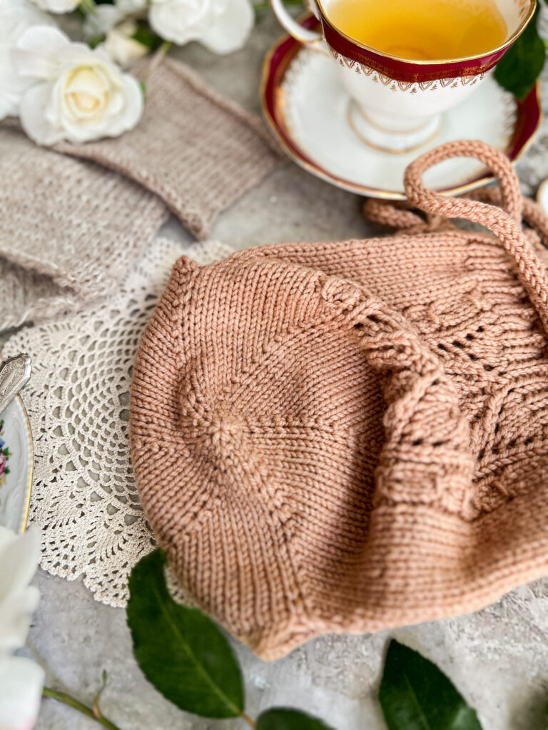 A close-up on the bottom of a tea cozy with radiating KFB increases. In the background, slightly blurred, are a teacup full of chamomile, some white roses, and three tan swatches.