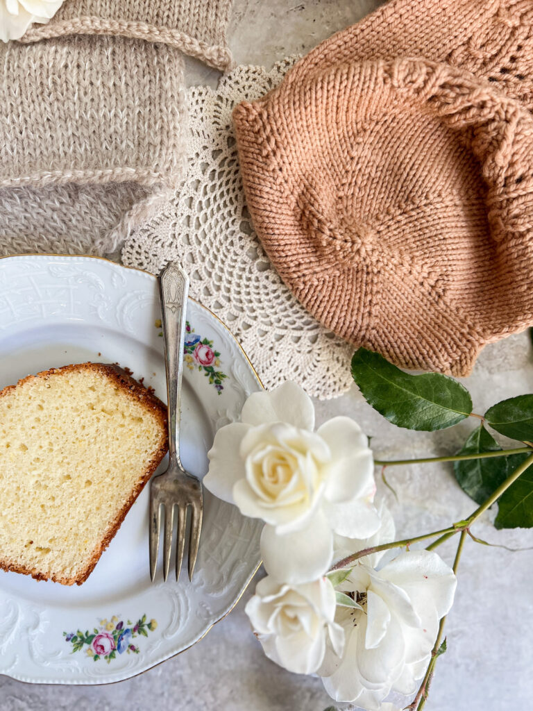 A flatlay photo showing the bottom of a tea cozy. The tea cozy has radiating spokes, which show how the KFB in knitting can be used as a decorative element. To the left of the image is a vintage floral plate with a slice of lemon yogurt cake on it, along with three tan swatches.