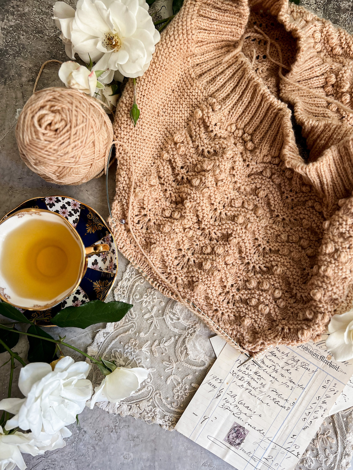 A flatlay photograph of a sweater vest in progress. It's tan and has lots of bobbles with rippling garter stitch. It's surrounded by white roses, a teacup full of chamomile, a lace handkerchief, and some antique paper ephemera.