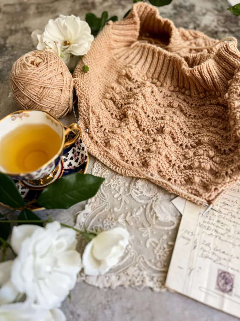 A tan sweater vest in progress with lots of rippling garter stitch and bobbles. It rests on top of an antique handkerchief and some paper ephemera, surrounded by white roses and a teacup full of chamomile.