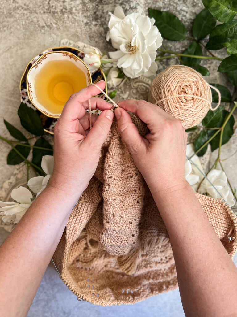 A white woman's hands (mine!) hold a tan knitting project and prepare to work a purl stitch. I am knitting in the continental style with the working yarn tensioned over my left index finger. in the background are a teacup full of chamomile tea and some white roses.