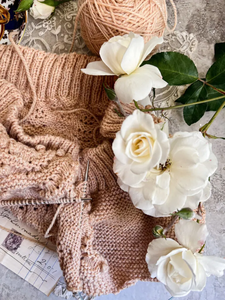 A few white iceberg roses rest against a knitting project in progress. The project is knit in tan wool. you can see the thick ribbing at the bottom of the project, along with lots of garter stitch and some bobbles.