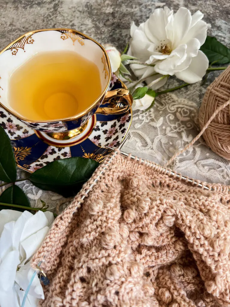 A close-up on the needles of a knitting project resting against the saucer of a blue floral teacup. The knitting project is a warm tan color and features bobbles and garter stitch.