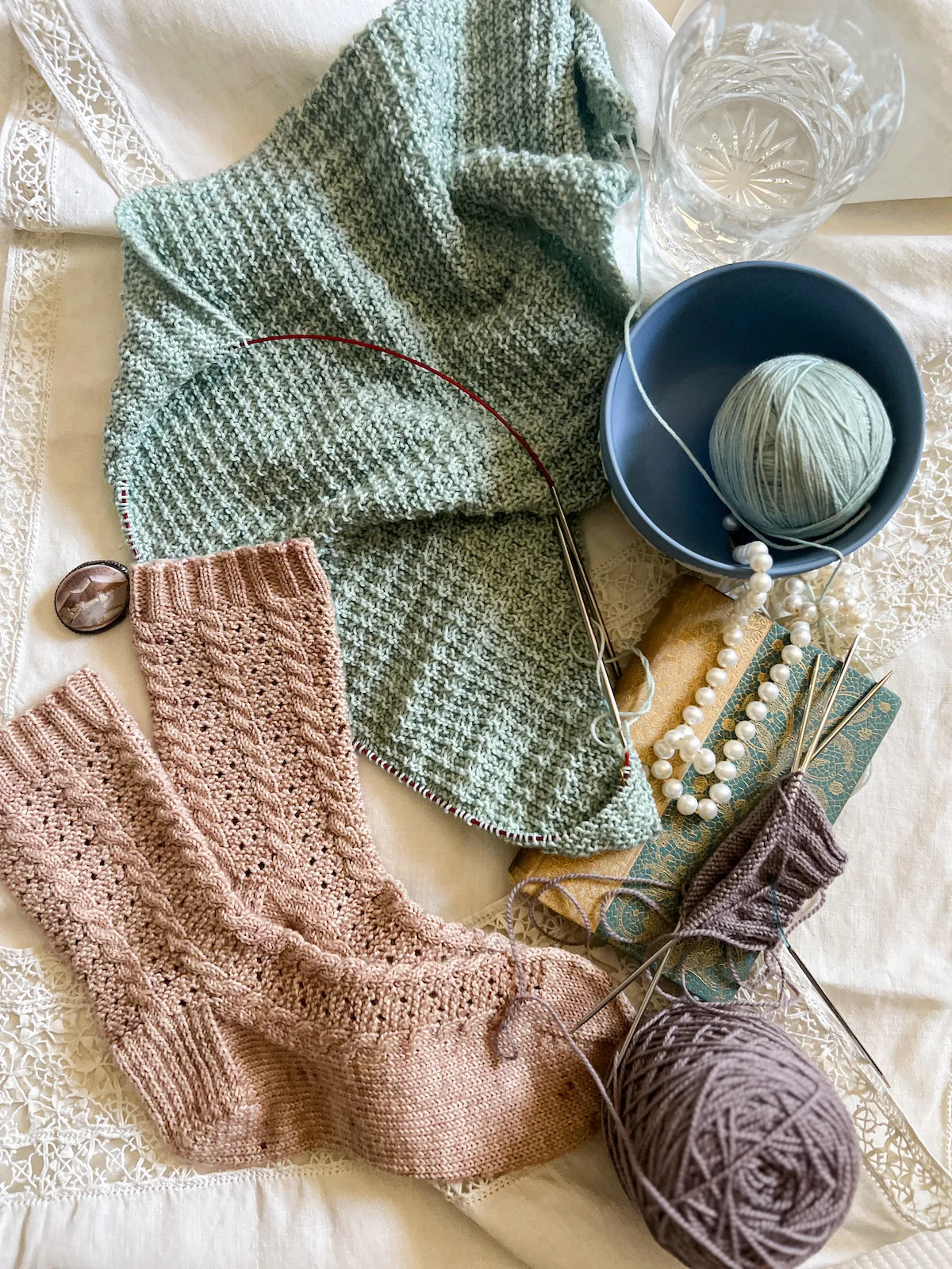A flatlay photo of a seafoam green knit shawl in progress, a finished pair of pink knit socks, and a barely started pair of light purple knit socks laid out on an antique table cloth with thick lace trim. Also in the image is an antique book with a gold and turquoise cover, a light blue Wedgwood bowl holding a ball of seafoam green yarn, a string of pearls, a crystal glass filled with water (it's very hot today), and a small carved shell brooch.