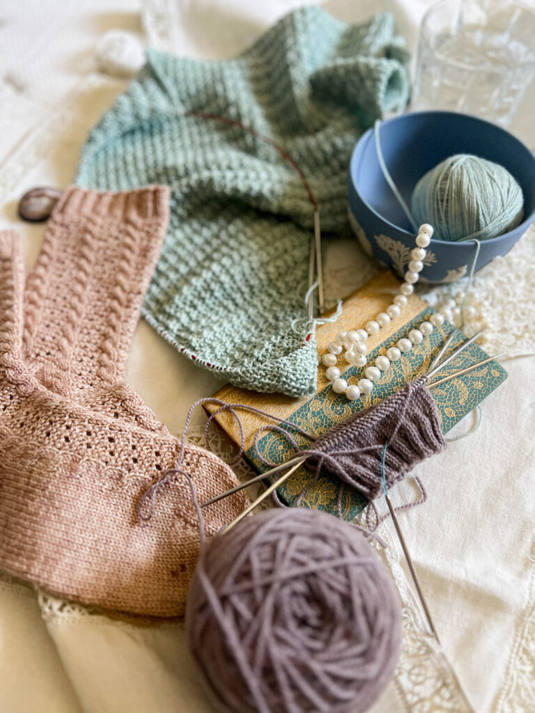 A side angle view of a pile of knit items: a seafoam green shawl in progress, some finished pink socks, and a barely started light purple sock. The shawl is at the top of the image, the pink socks are to the left, and the purple sock takes up the center right. There's also an antique book, some pearls, and a Wedgwood bowl with a ball of yarn in it.