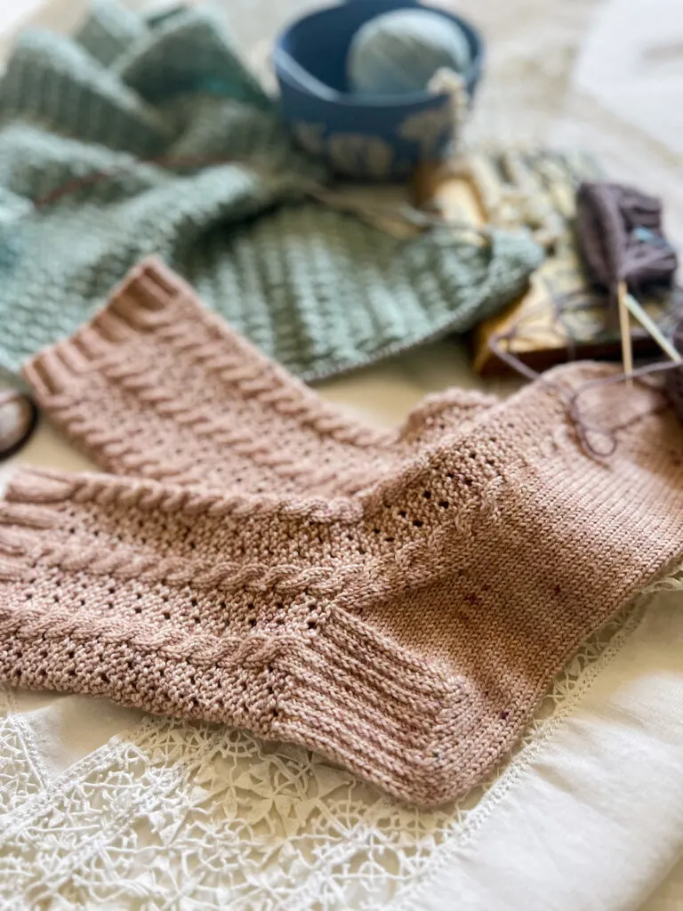 A pair of pink knit socks fills the bottom 2/3 of the image. Blurred in the background are some other pastel knits and a light blue Wedgwood bowl holding a ball of yarn.