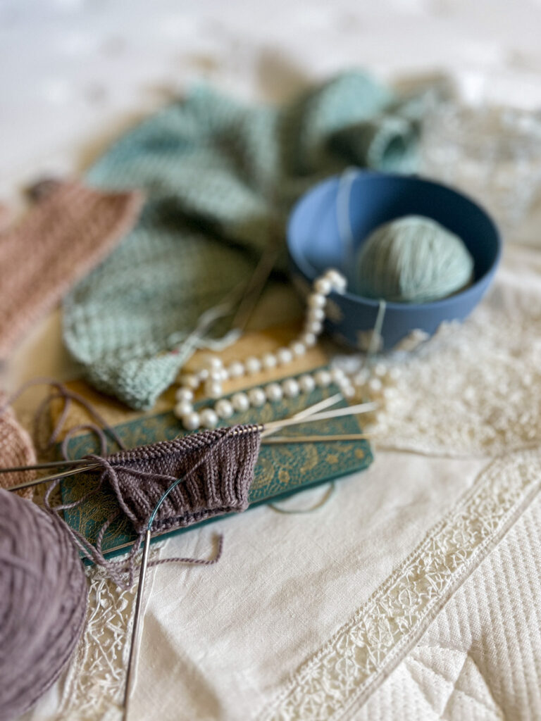 A tightly focused image narrows in on a light purple sock in progress on a pair of Addi Flexiflip needles at the bottom left of the photo. Blurred in the background are other pastel knits, a string of pearls, and a light blue Wedgwood bowl holding a ball of yarn.